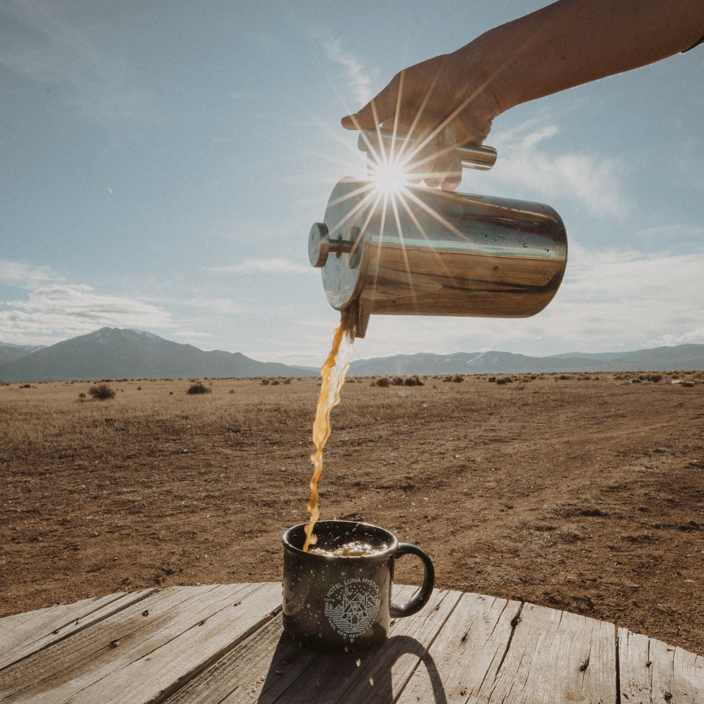 Pouring coffee from a French press into a black mug on a wooden table, with a scenic desert landscape and mountains in the background, showcasing outdoor brewing at Cottonwood Coffee.